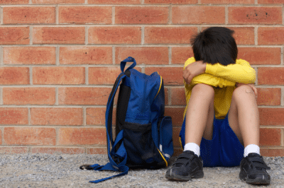 sad boy sitting against a brick wall with backpack