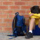 sad boy sitting against a brick wall with backpack