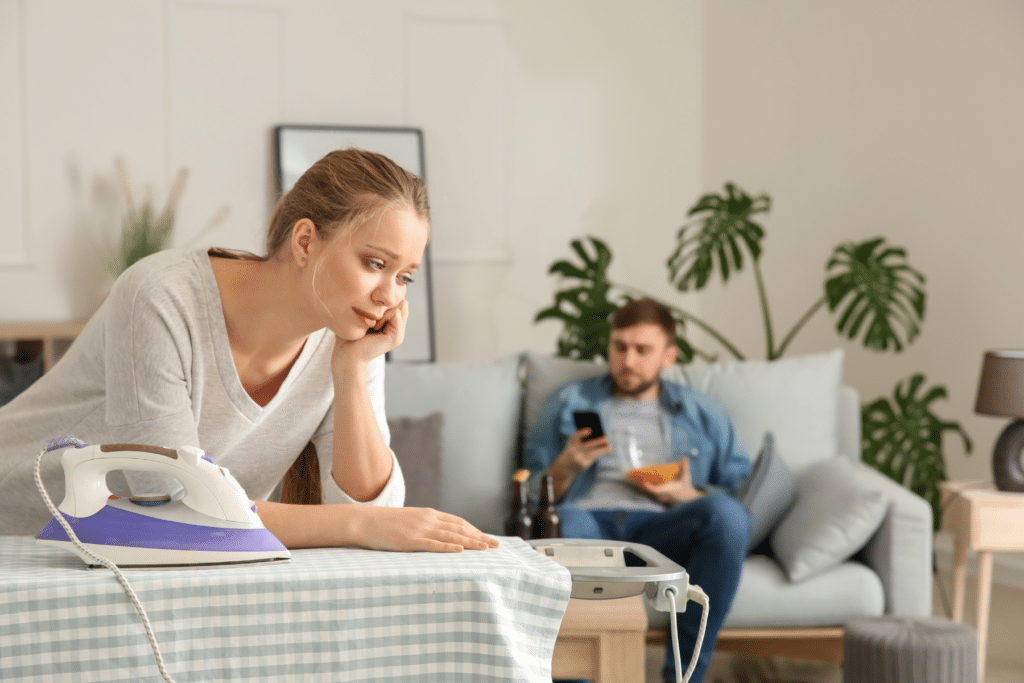 woman looking sad leaning on an ironing board and a man sitting on a couch eating snacks and looking at his smart phone