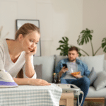 woman looking sad leaning on an ironing board and a man sitting on a couch eating snacks and looking at his smart phone