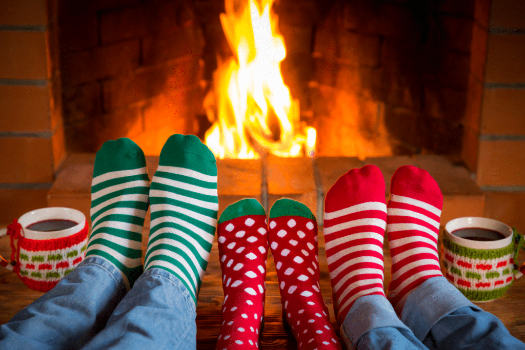 Three sets of feet in Christmas socks in front of a fireplace with two mugs of coffee