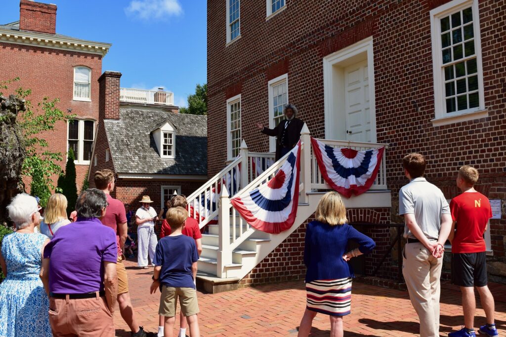 A crowd of people gather outside a historic building with red, white, and blue banners decorating the rails of the stairs.