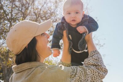 a young, white mother with dark hair holds her baby up in the air with the shining sun in the sky above