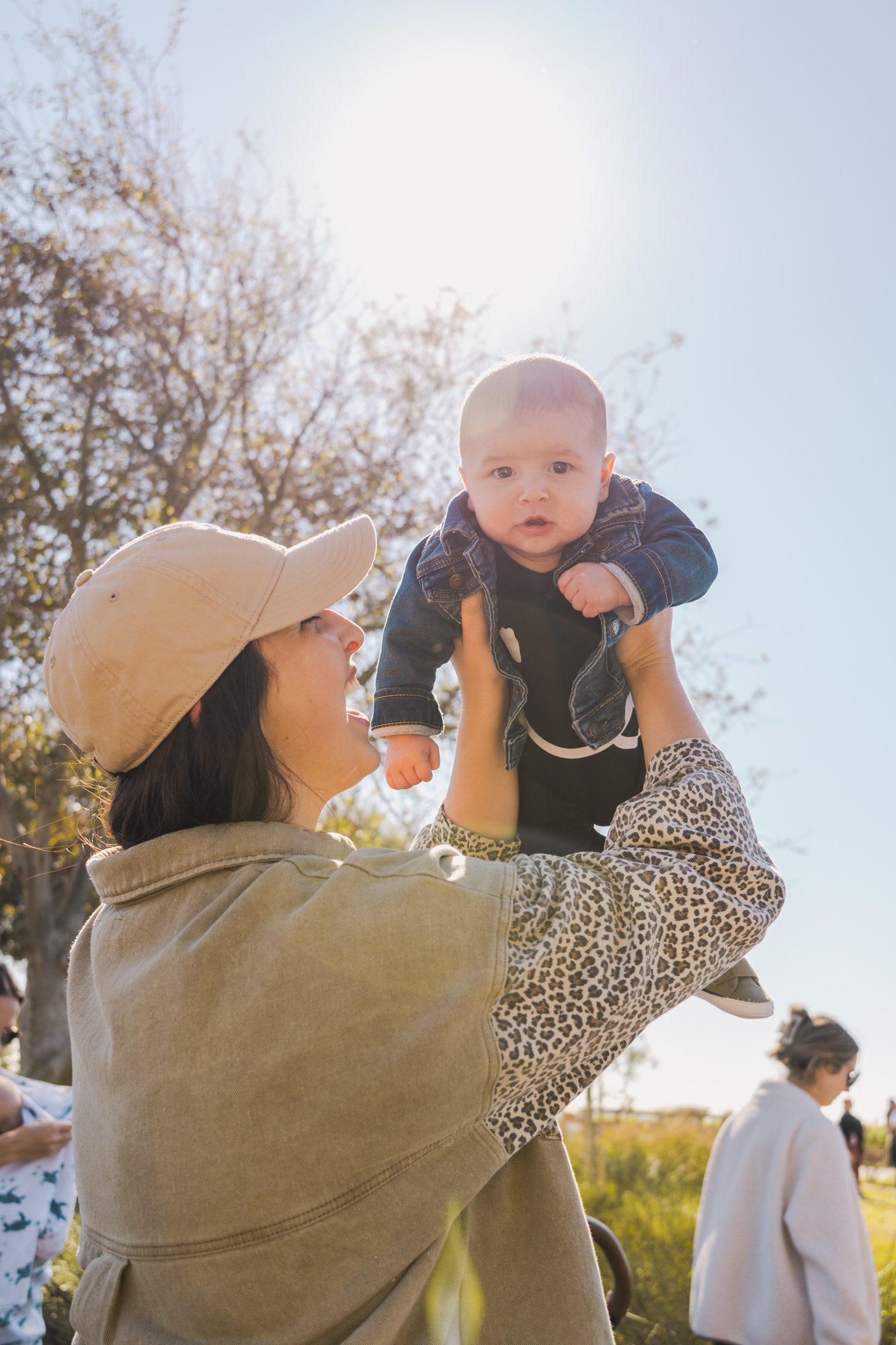 a young, white mother with dark hair holds her baby up in the air with the shining sun in the sky above