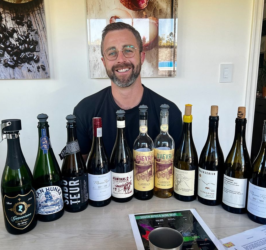 A man, Peter Andrews, sits smiling in front of several bottles of South African wine