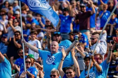 a crowd of people wearing blue cheer in the stands at the Annapolis Blues FC Mid-Atlantic Conference Championship.