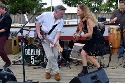a white man in a white shirt plays an electric guitar and a white woman in a black tank top and skirt plays a fiddle