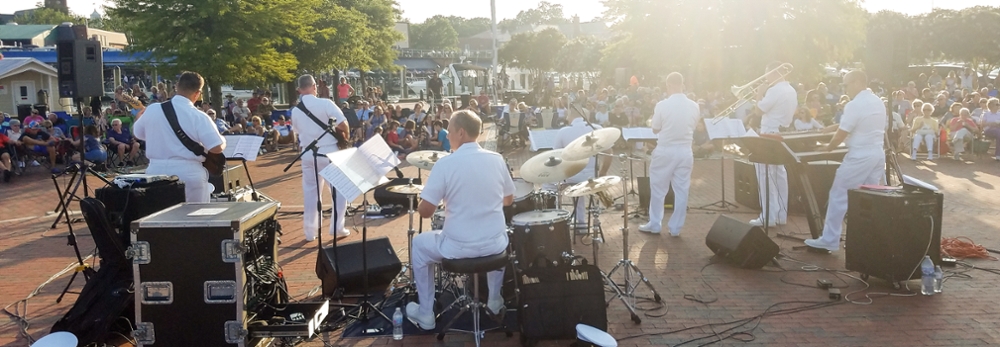 a band in white navy uniforms performs outside, captured from behind