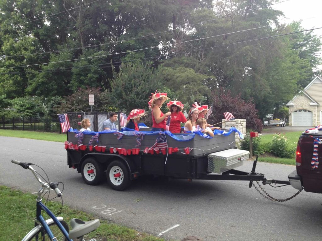A group of people sit and stand in a float decorated for the Cape St. Claire Fourth of July Parade that's moving down the street.