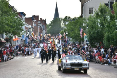 A white car decorated with small American flags rolls through downtown on the road in the Annapolis Independence Day Parade with men in uniform walking behind it.